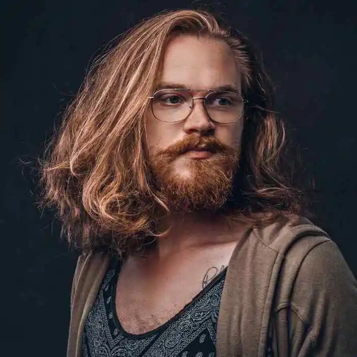 15 Best Wavy Hairstyles for Men 2 A man showcasing men wavy hairstyles with long, wavy brown hair stands against a dark backdrop, wearing a patterned shirt and a loose sweater.