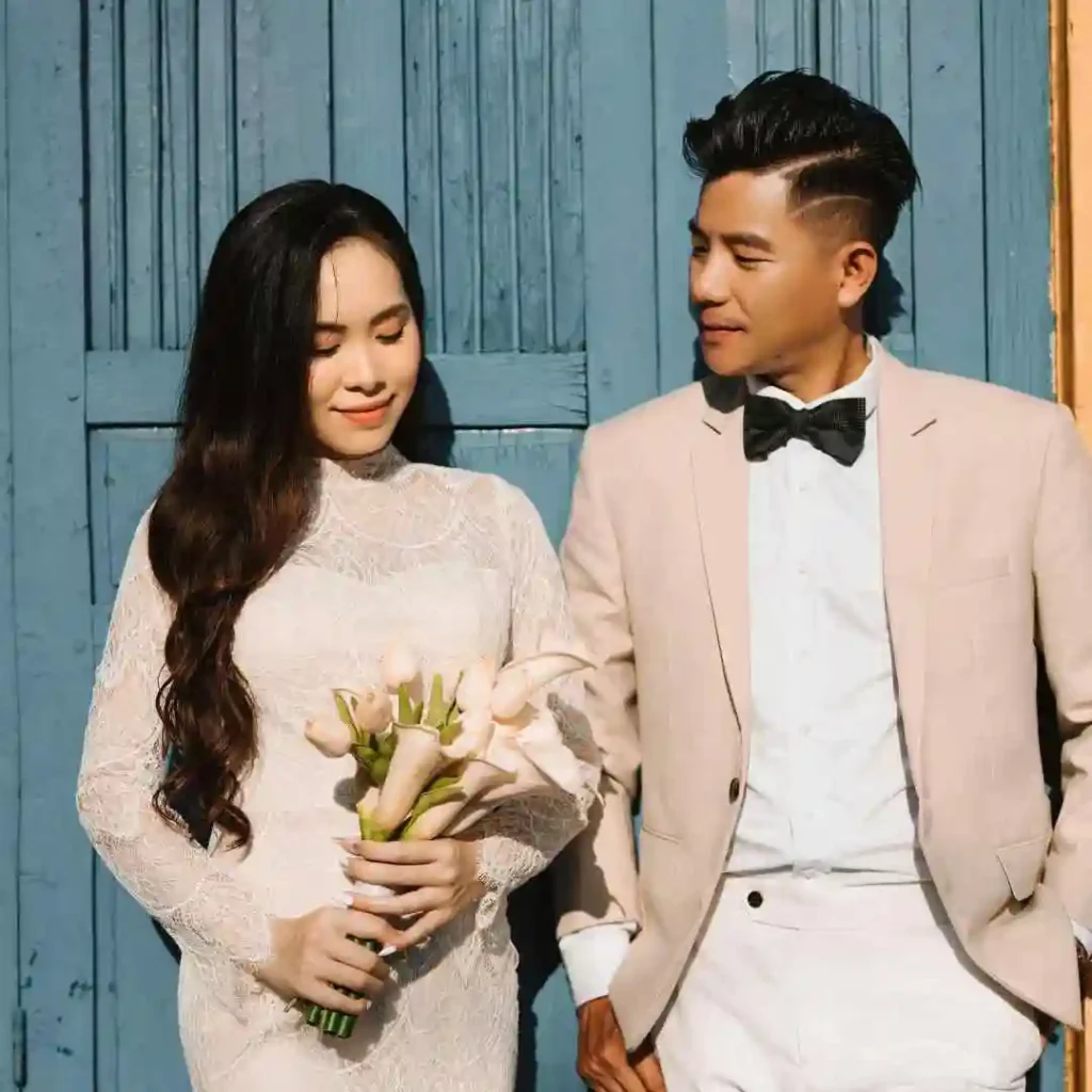 27 Wedding Hairstyles for Men That Can Elevate Your Style 7 A couple stands together in front of a blue wooden door, the woman holds a bouquet of white flowers, both dressed elegantly; the man showcases a stylish wedding hairstyle for men.