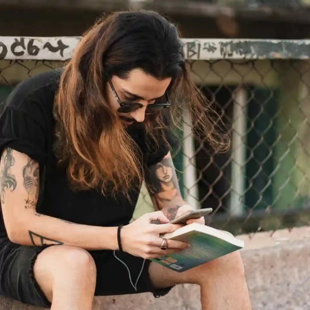 Long Hair Styling for Men.. Creative Hairstyles And Ideas 2 A person showcasing Long Hair Styling for Men, with long hair and tattoos, sits on a ledge holding a smartphone and a book, set against a backdrop of lush greenery.