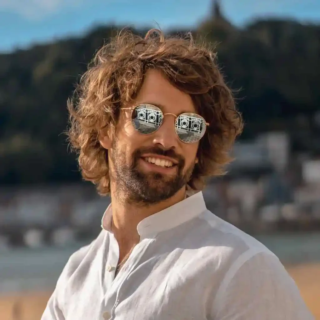 A man with curly hair and glasses wears a white collared shirt, standing against a scenic outdoor backdrop with hills and a body of water, showcasing men hairstyles with glasses.