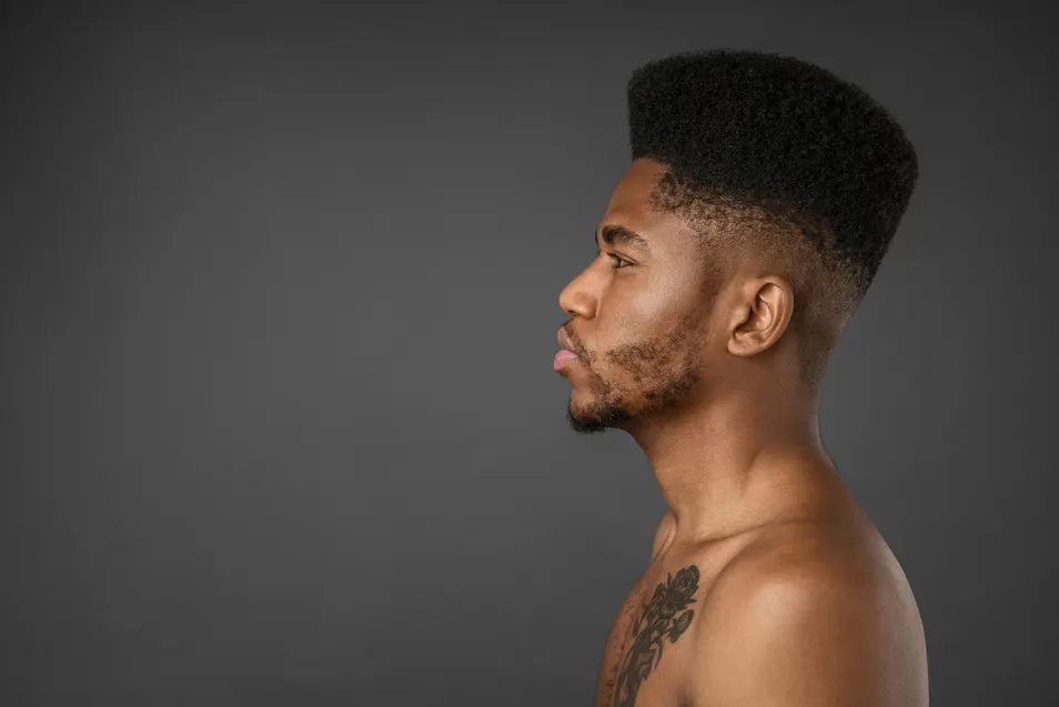 "Profile of a shirtless person with a tall, textured fade hairstyle for men and a visible tattoo on their upper arm, set against a dark background.