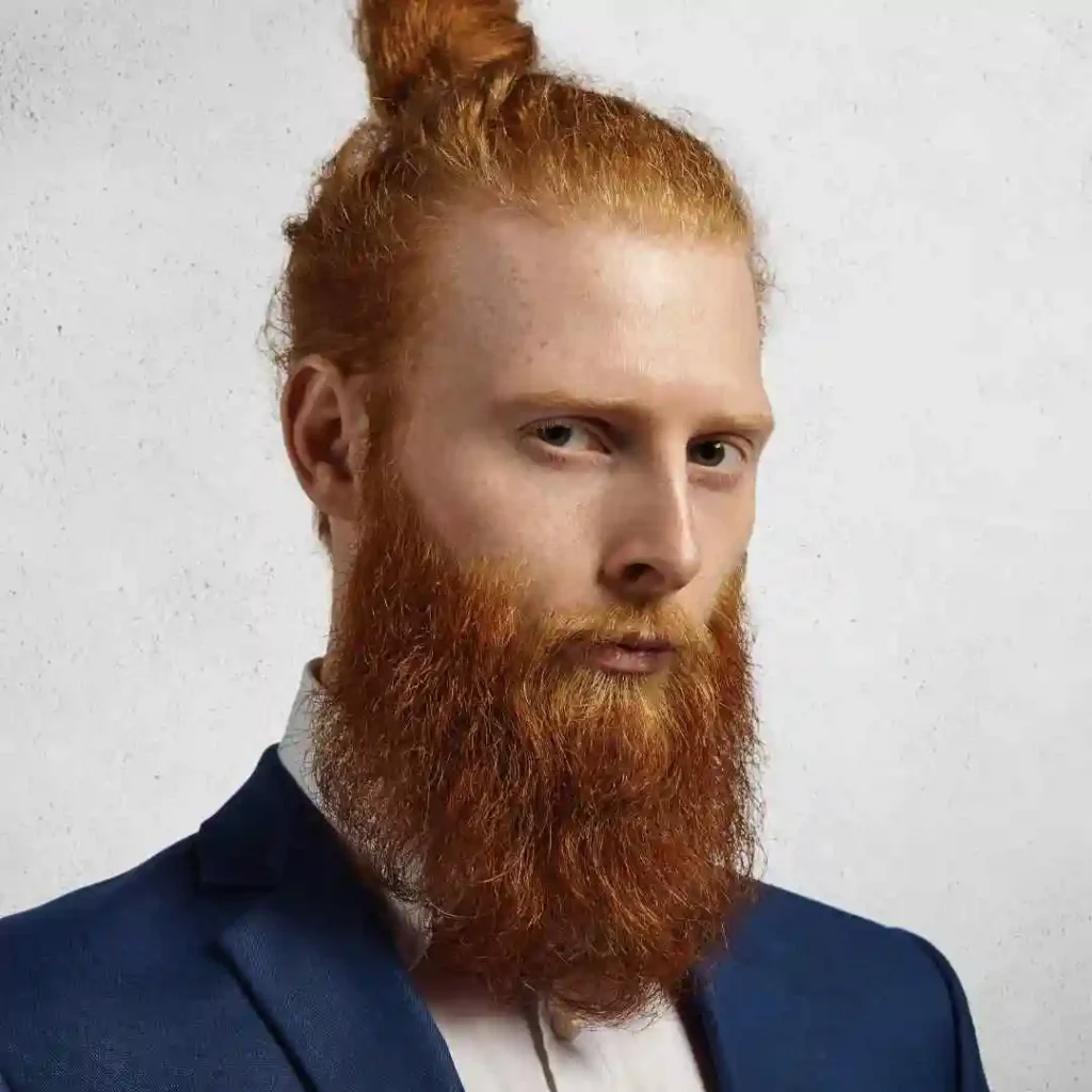 A man with a reddish beard and long hair styled in a bun, showcasing long hair styling for men, wearing a dark suit and white shirt, posed against a plain light background.