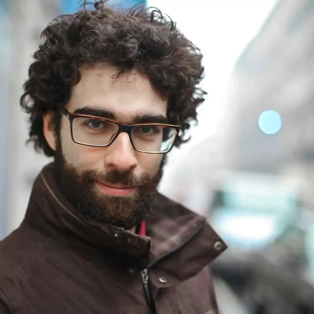 A person with curly hair wearing glasses and a brown jacket showcases stylish men hairstyles with glasses while standing on a street with a blurred cityscape in the background.
