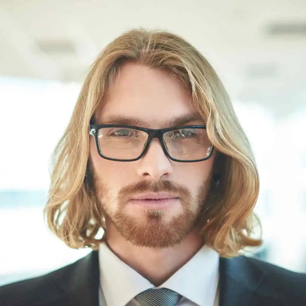 A man with long, wavy blonde hair and glasses, dressed in a suit and tie, stands in a bright, modern office environment showcasing stylish men hairstyles with glasses.