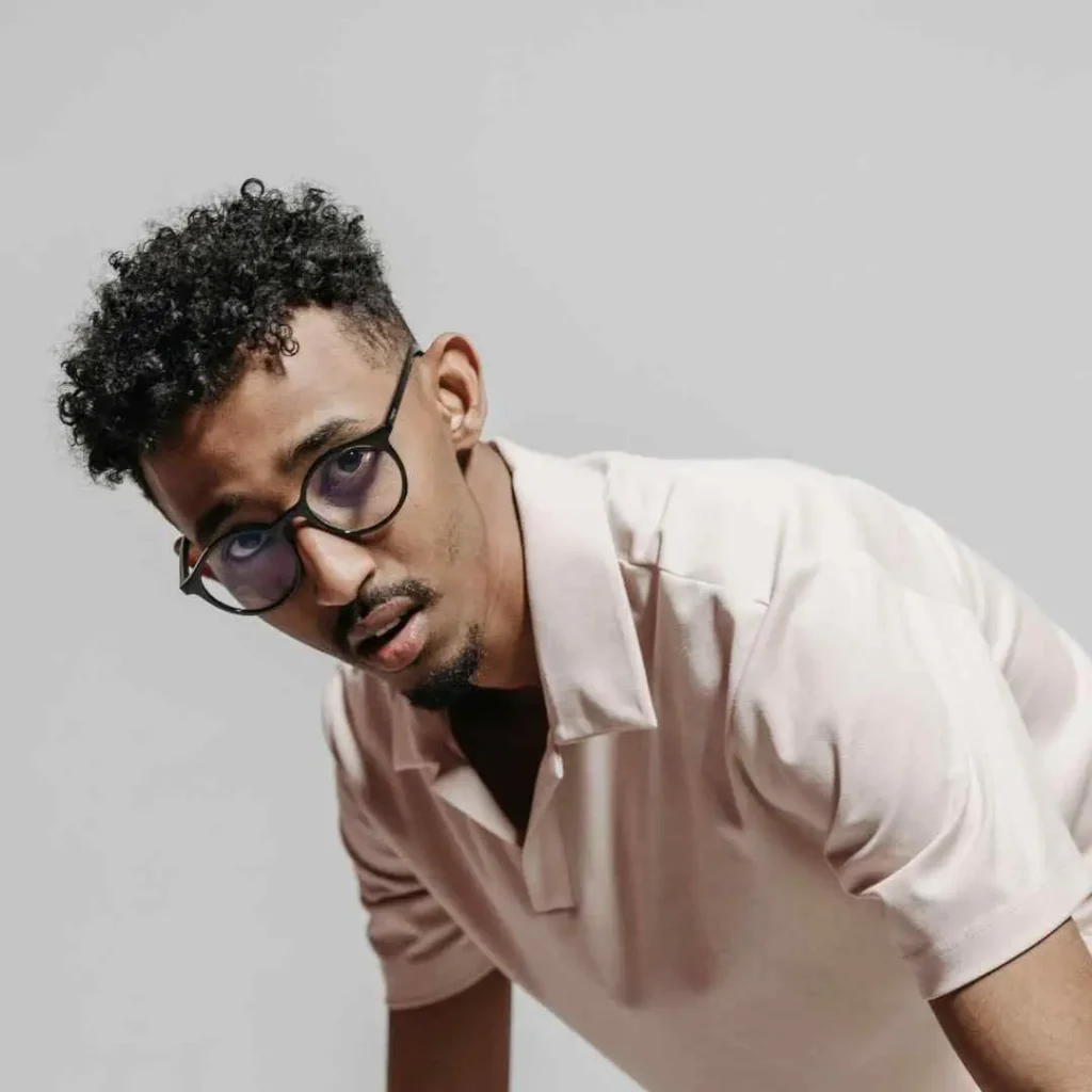 A person with stylish men hairstyles with glasses leans forward, wearing a light pink polo shirt over a dark t-shirt, against a soft gray background.