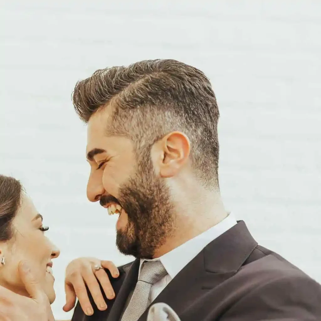 A man in a black suit showcasing a stylish wedding hairstyle for men and a woman in a dress share a playful moment against a light, textured background.
