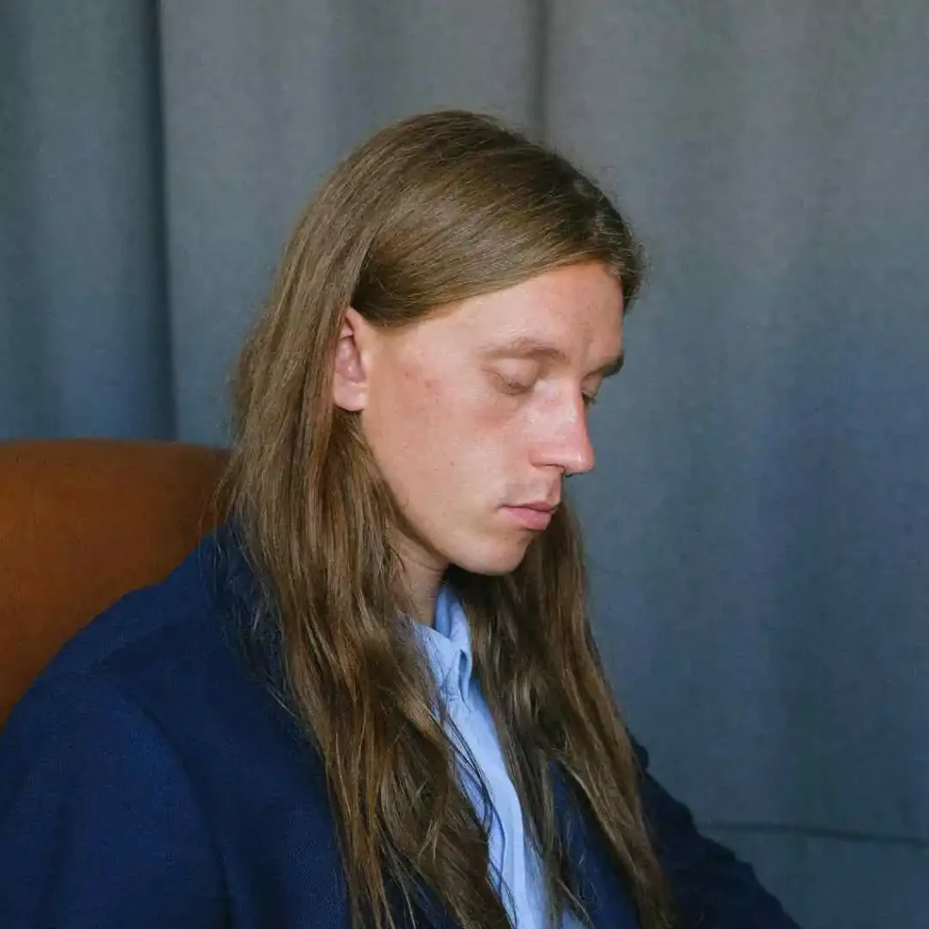 A person with long, brown hair styled for men, seated in a chair, wearing a dark jacket and blue shirt, against a grey backdrop.