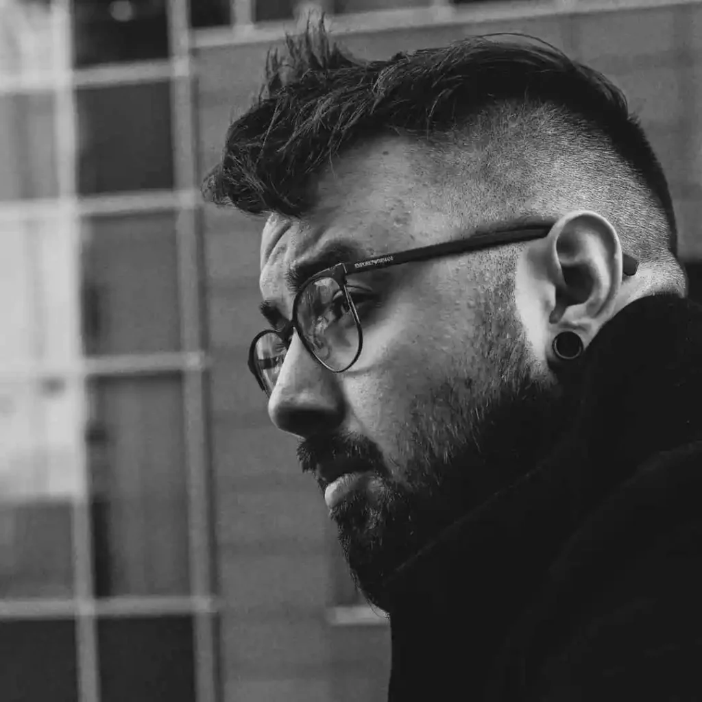 A profile of a person showcasing men hairstyles with glasses, featuring a stylish haircut, wearing glasses and earrings, against a backdrop of blurred architectural lines.