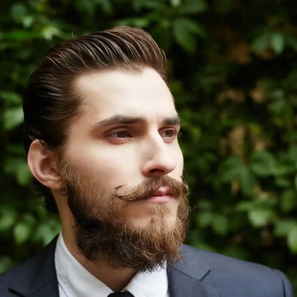 A man with slicked-back hair and a neatly groomed beard wears a suit in front of a lush green background, showcasing a perfect wedding hairstyle for men.
