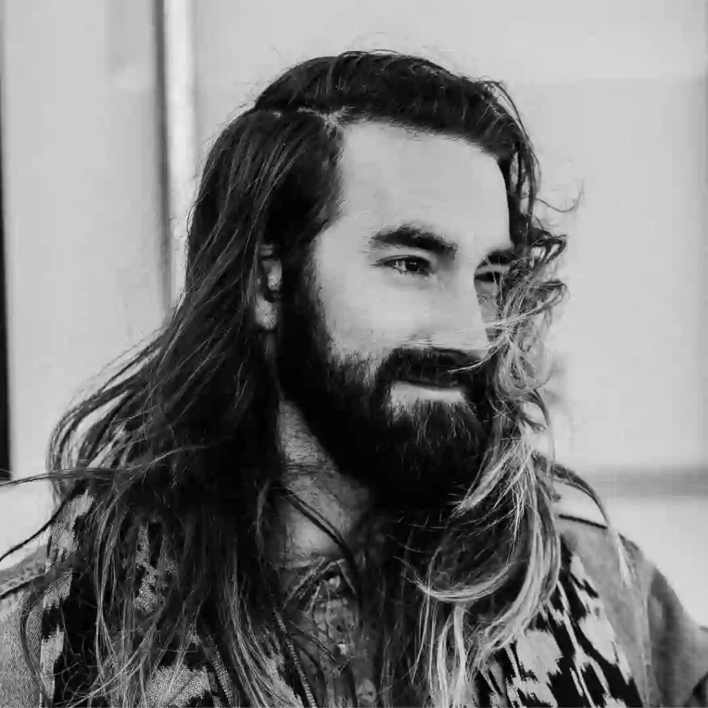 A person with long, wavy hair styled for men is wearing a patterned garment, set against a neutral background. The image is in black and white.