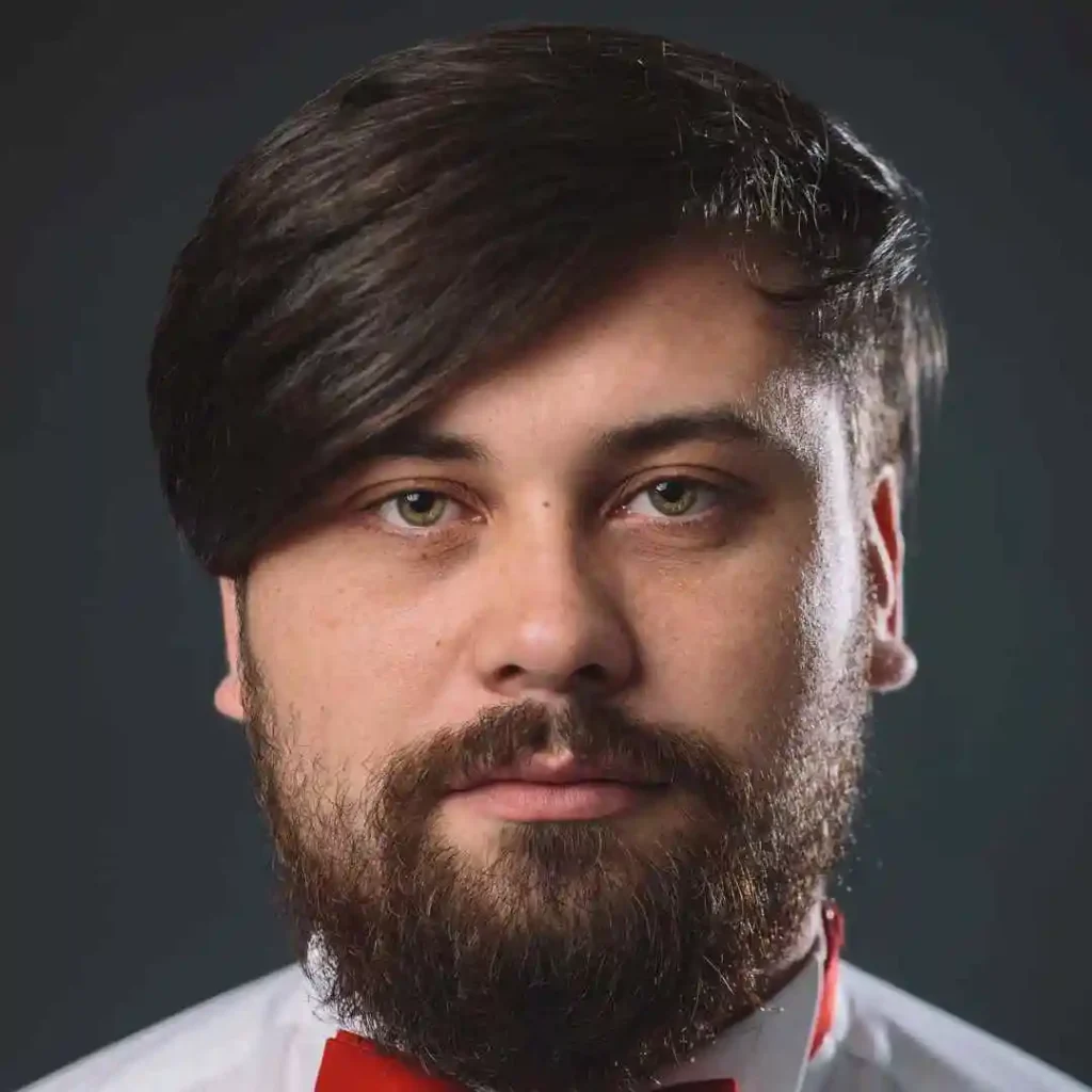 A man with dark hair and a beard is wearing a white shirt and red bow tie against a dark background, showcasing a formal appearance with a stylish wedding hairstyle for men.