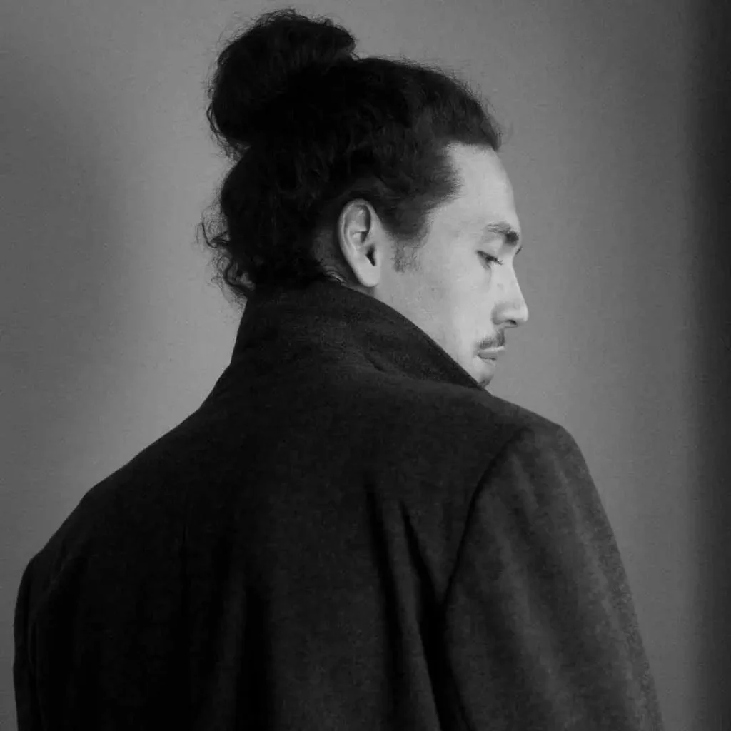 A person with long, curly hair styled in a bun, showcasing long hair styling for men, wears a dark coat, facing away against a soft gray background. Black and white photo.