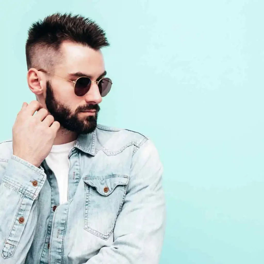 A person with short hair wearing glasses and a denim jacket poses against a light blue background, resting a hand on their chin thoughtfully, showcasing men hairstyles with glasses.