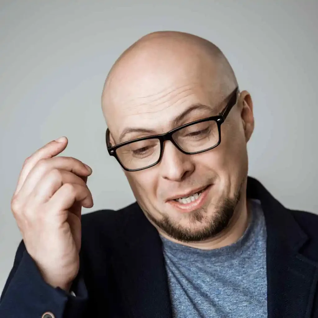 A bald man with stylish men hairstyles with glasses, wearing a grey shirt and dark blazer, gestures with one hand against a soft grey background.