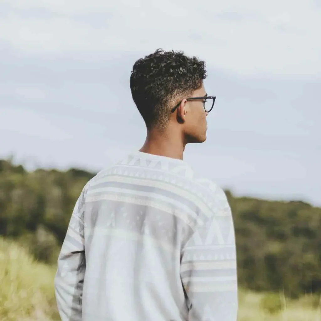A person wearing a patterned sweater and glasses stands on a hillside, showcasing men hairstyles with glasses, gazing into the distance under a cloudy sky.