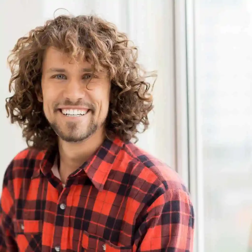 15 Best Wavy Hairstyles for Men 3 A person showcasing men wavy hairstyles, with curly hair wearing a red and black plaid shirt, sitting by a window with soft natural light.