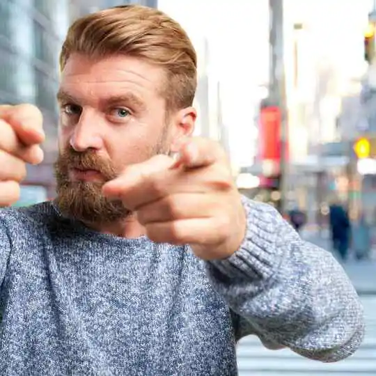 Cool And Breezy Summer Hairstyles for Men 3 A person in a gray sweater with a trendy summer hairstyle for men stands outdoors, playfully pointing fingers at the camera against a bustling city background.