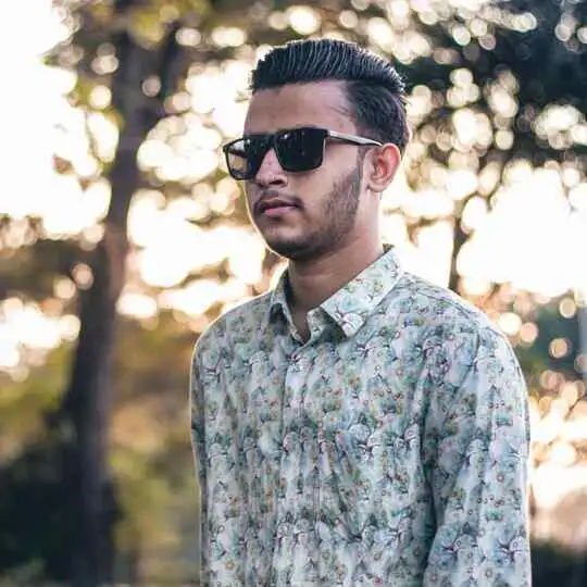 A person with stylish men hairstyles and glasses, wearing a floral shirt, stands outdoors surrounded by trees and soft golden light, creating a serene atmosphere.