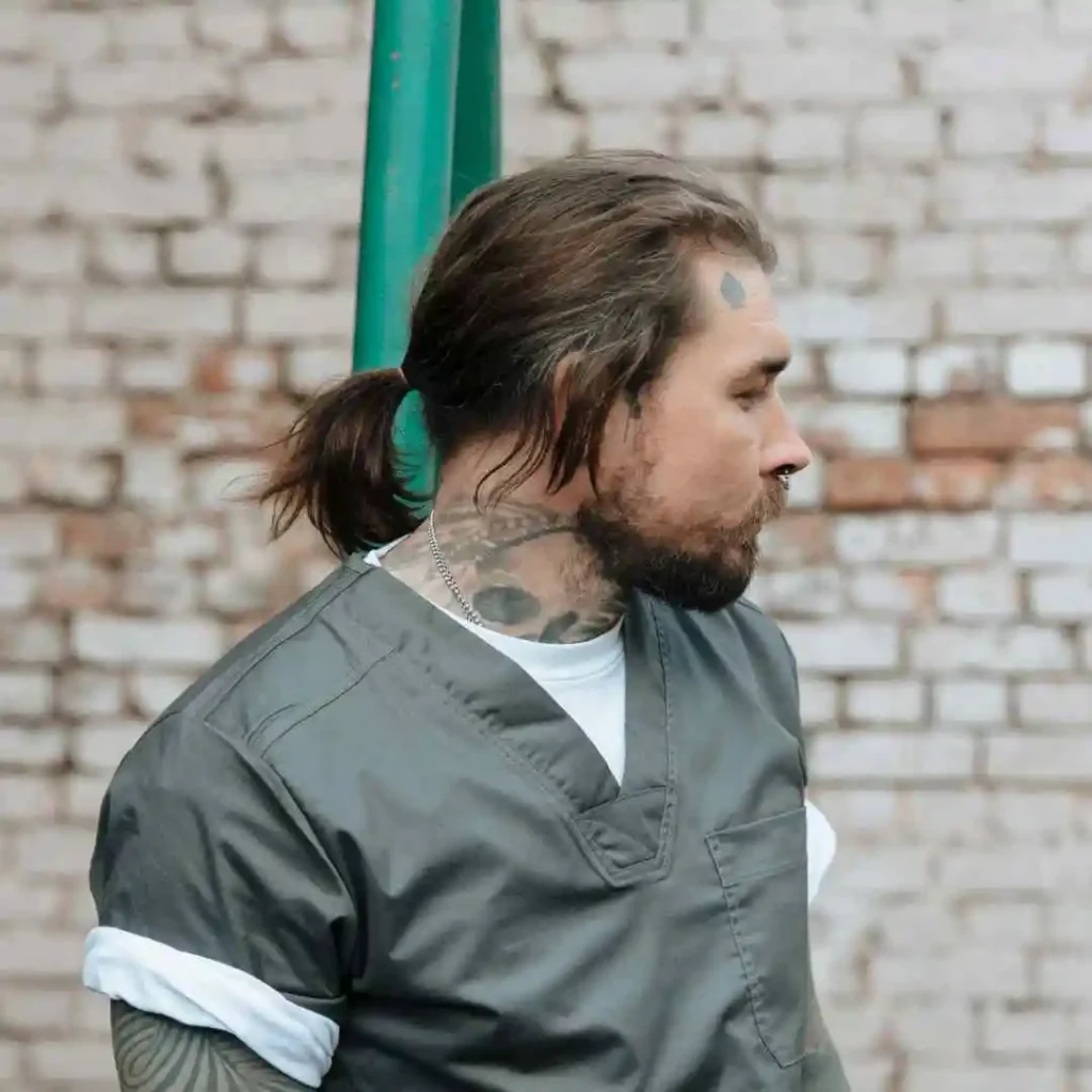 Long Hair Styling for Men.. Creative Hairstyles And Ideas 3 A person showcasing long hair styling for men, with their hair tied back, wearing a gray medical uniform, standing against a rustic brick wall with visible tattoos.