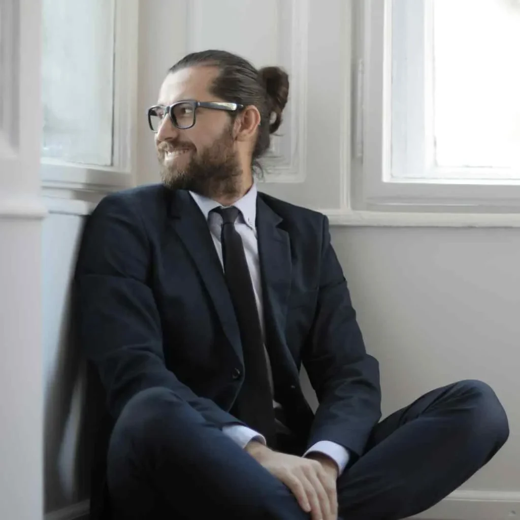 A man in a tailored suit and tie with men hairstyles with glasses sits cross-legged against a wall near a window, exuding a sense of contemplation and professionalism.