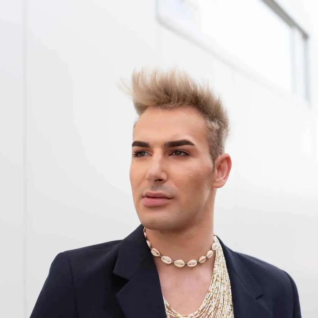 A person wearing a stylish black blazer and layered shell necklaces stands against a light-colored background, showcasing a wedding hairstyle for men.