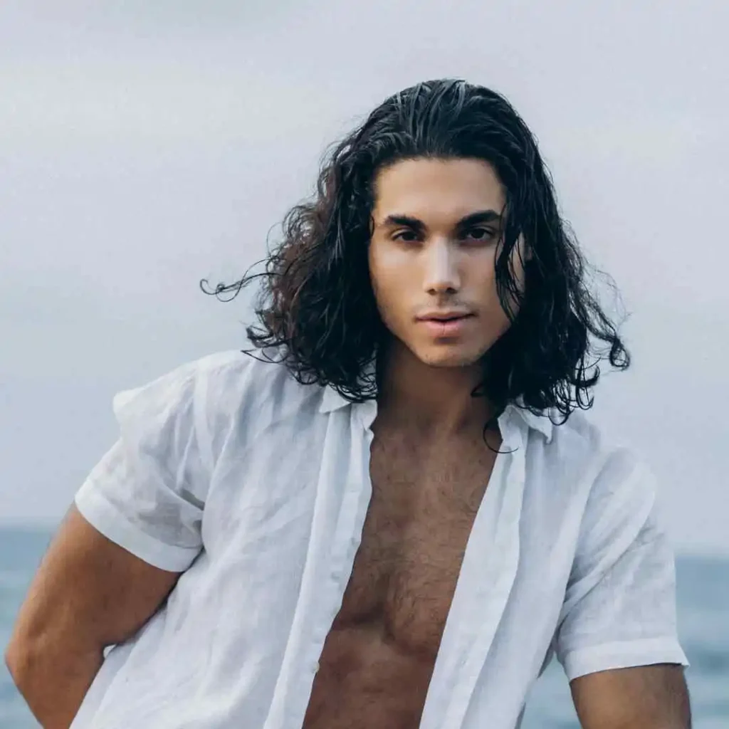 A person showcasing Long Hair Styling for Men with long, wavy hair poses by the ocean, wearing an open white shirt that reveals their torso.