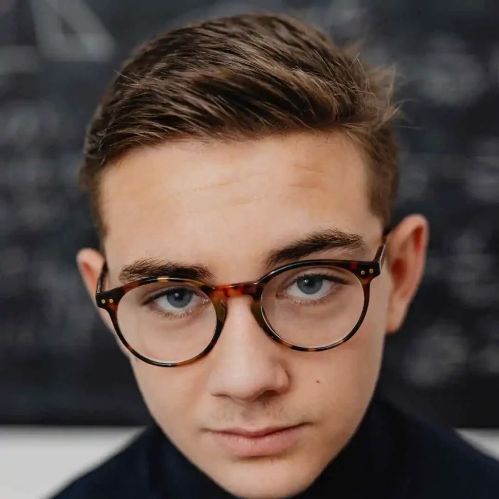 A young person with styled brown hair and glasses, showcasing men hairstyles with glasses, is wearing earbuds, set against a blurred chalkboard background with faint writing.