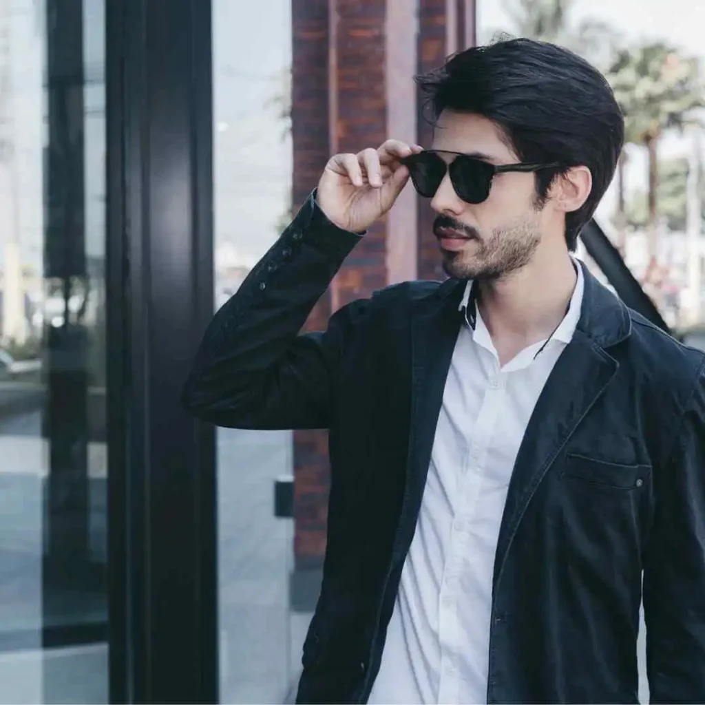 A young man with stylish men hairstyles with glasses, wearing a black jacket and white shirt, stands outdoors adjusting his sunglasses in front of a modern building.