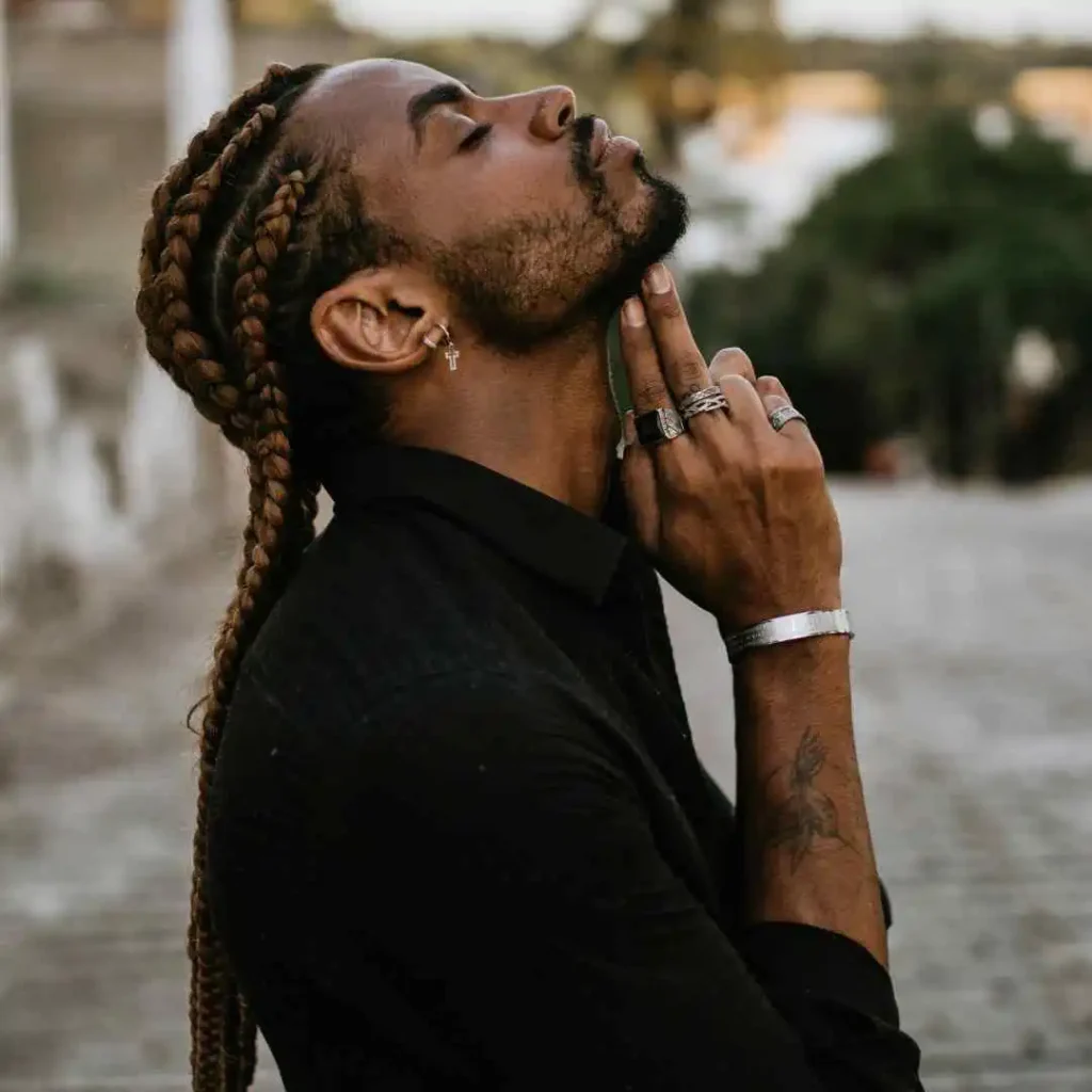A person with braided hair showcasing long hair styling for men, wearing multiple rings and a silver bracelet, looking contemplatively towards the sky against a blurred outdoor background.