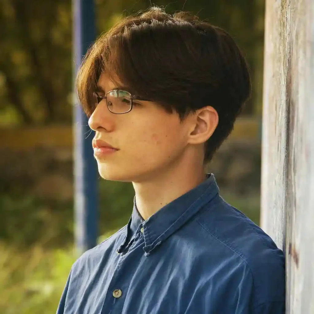 A young person with stylish men hairstyles with glasses stands against a textured wall, wearing a blue button-up shirt, with greenery softly blurred in the background.