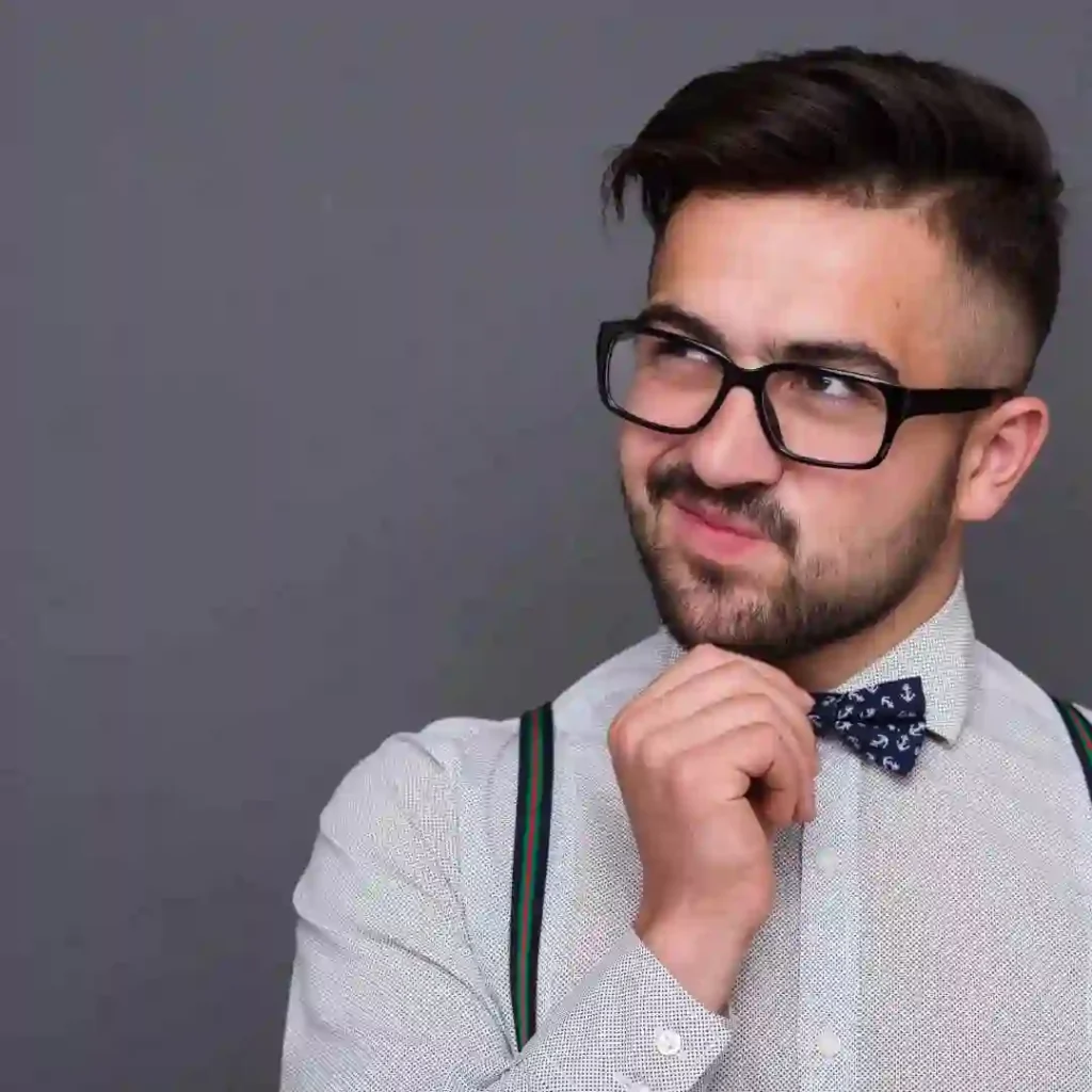 A well-dressed individual with stylish men hairstyles and glasses adjusts a blue anchor-patterned bow tie, wearing a button-up shirt with suspenders against a gray background.