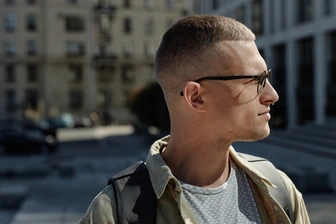 A young man showcasing trendy men hairstyles with glasses, wearing sunglasses and a casual outfit, standing outdoors with modern architecture and parked cars in the background