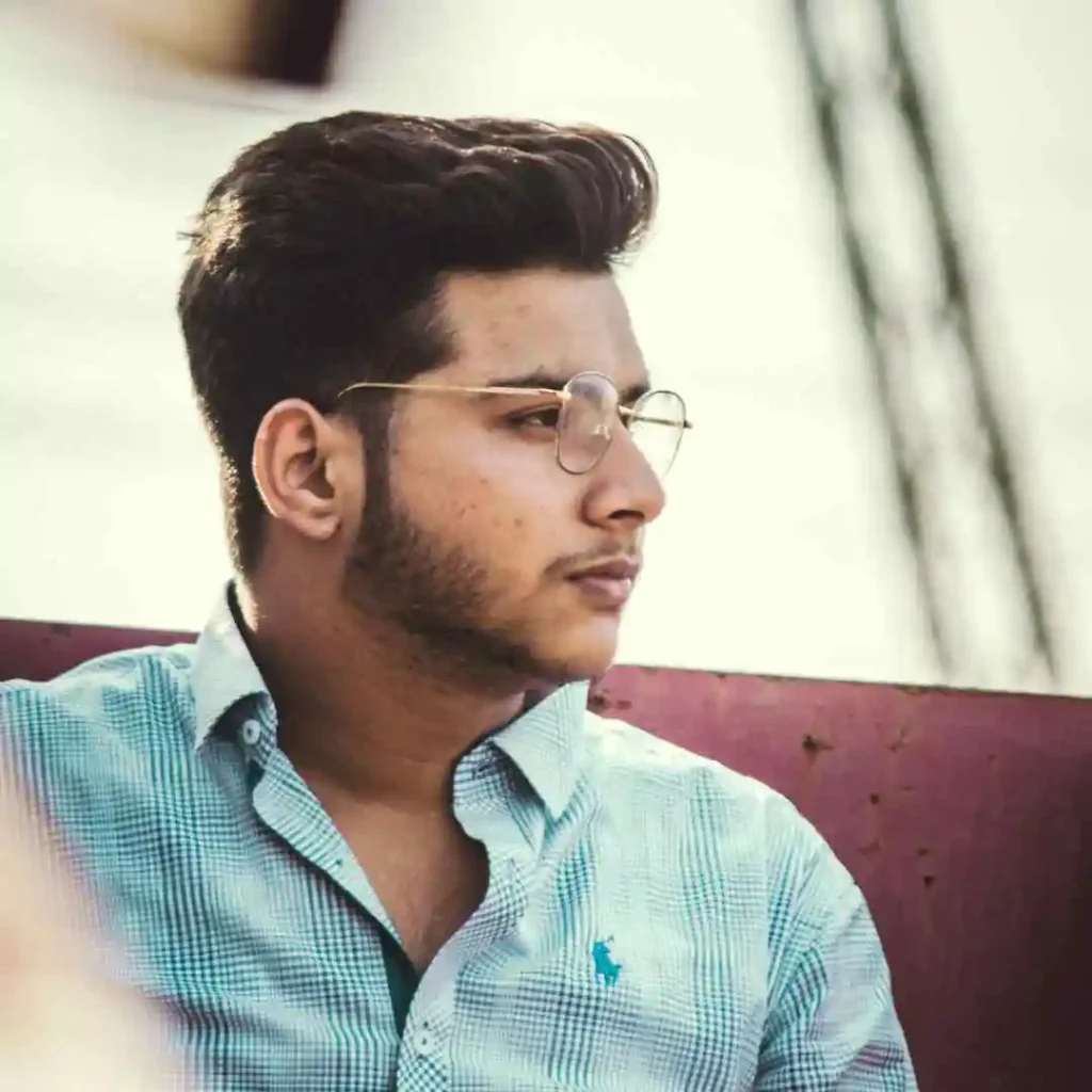 A young man showcasing trendy men hairstyles with glasses, wearing a light blue checkered shirt and sitting against a maroon backdrop, exuding a relaxed vibe.