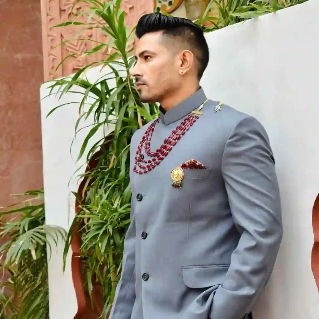 27 Wedding Hairstyles for Men That Can Elevate Your Style 6 A man showcasing a Wedding Hairstyle For Men, dressed in a stylish grey suit adorned with red jewelry and a decorative pin, standing against a backdrop of green plants.