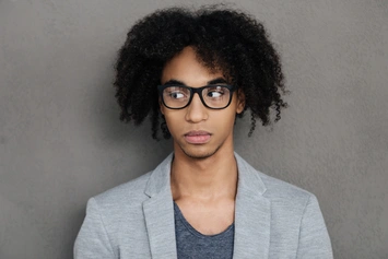 A person showcasing Men Hairstyles with Glasses, featuring curly hair, wearing a grey blazer and a blue shirt, standing against a plain gray background