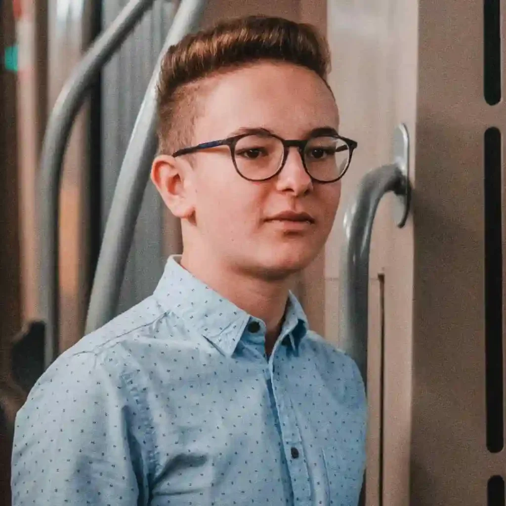 A young man showcasing trendy men hairstyles with glasses, wearing a light blue patterned shirt, stands next to a railing in an indoor setting with warm lighting.