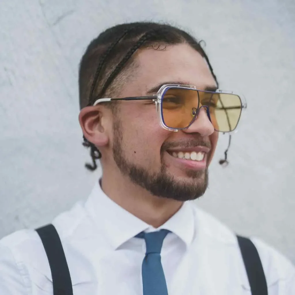 A person showcasing men hairstyles with glasses, featuring braided hair, wearing a white shirt and blue tie, posed against a light gray wall.