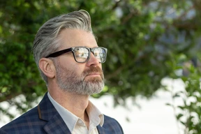 A man with silver hair showcasing trendy men hairstyles with glasses stands outdoors, wearing a stylish plaid suit against a backdrop of greenery.