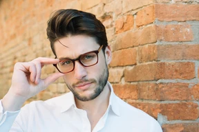 A man with stylish men hairstyles with glasses, wearing a white shirt, poses against a rustic brick wall, with one hand touching his fingers near his face.