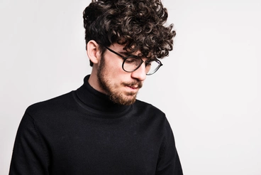A young person with curly hair wearing glasses and a black turtleneck, showcasing trendy men hairstyles with glasses, looking thoughtfully to the side against a plain white background.
