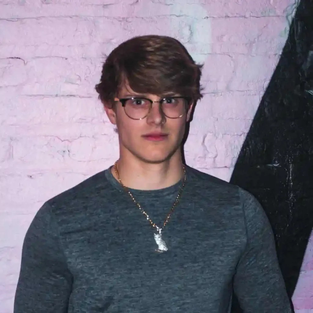 A person with stylish men hairstyles with glasses, wearing a gray long-sleeve shirt and a silver necklace, stands against a textured pink wall with black geometric shapes.