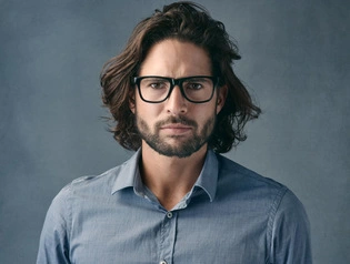 A person with long, wavy hair and glasses, showcasing men hairstyles with glasses, wearing a light blue button-up shirt, standing against a textured gray background.