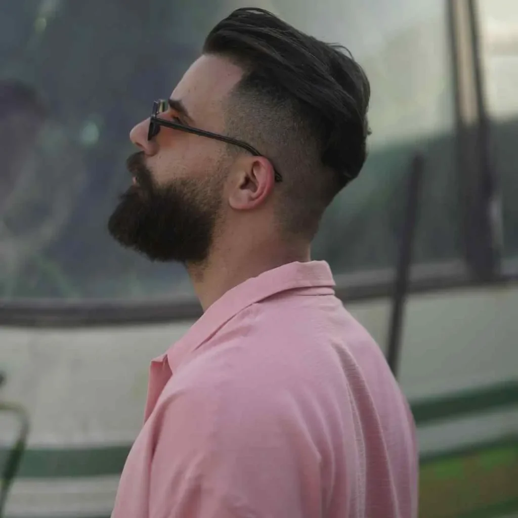 Cool And Breezy Summer Hairstyles for Men 5 A man showcasing one of the trendy summer hairstyles for men, with stylish hair and a beard, wearing a light pink shirt, standing against a blurred outdoor backdrop.