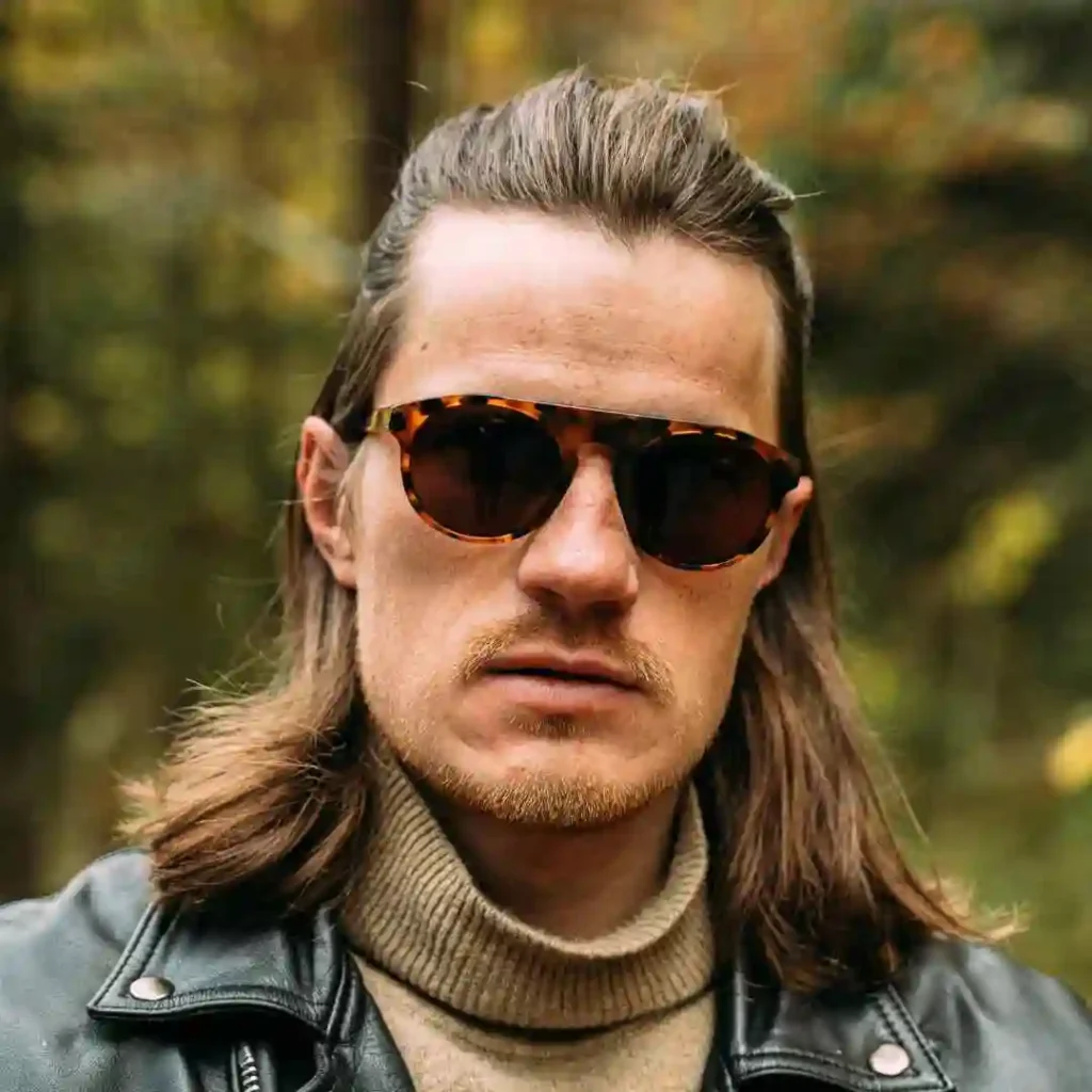 Long Hair Styling for Men.. Creative Hairstyles And Ideas 5 A person showcasing Long Hair Styling for Men, wearing a black leather jacket and a tan turtleneck, stands confidently in a vibrant, autumn forest.