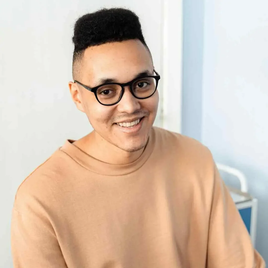 A person showcasing trendy men hairstyles with glasses, wearing a beige sweatshirt and sitting against a light blue background, with a soft focus on their features.