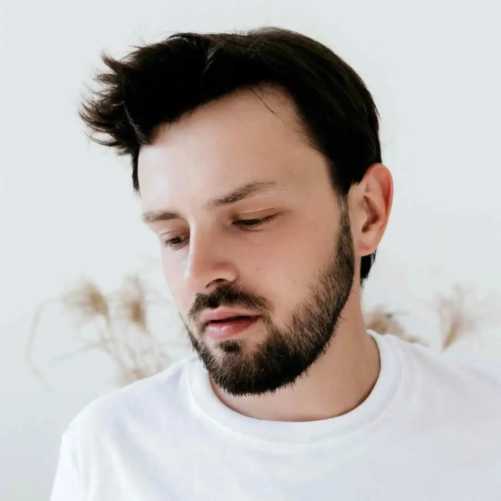 A person with dark, tousled hair showcasing a stylish Wedding Hairstyle For Men, wearing a plain white t-shirt, set against a soft, neutral-toned background with subtle plant decorations.