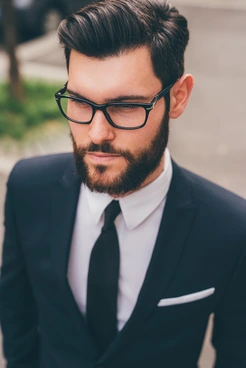 A man with stylish men hairstyles with glasses, dressed in a tailored black suit and white shirt with a slim tie, standing outdoors amidst greenery.