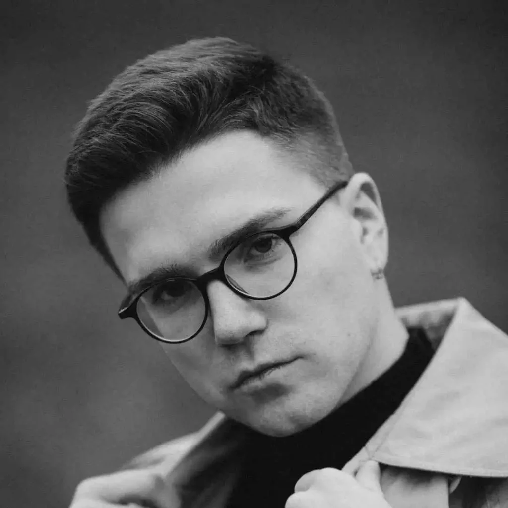 A person in a black and white photo, showcasing trendy men hairstyles with glasses, wearing a coat, and gazing sideways with a neutral expression against a blurred dark background.