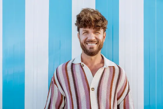 Cool And Breezy Summer Hairstyles for Men 6 A person showcasing Summer Hairstyles for Men stands in front of a blue and white striped wall, wearing a colorful, striped sweater with a buttoned collar.