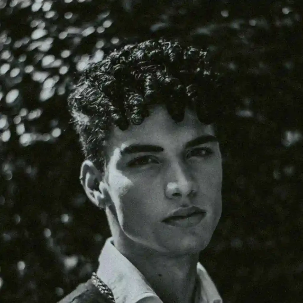A young man stands against a softly lit, blurred background, showcasing textured curly hair and a crisp shirt, demonstrating a stylish wedding hairstyle for men.